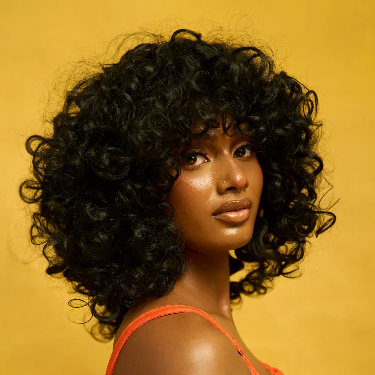 3/4 profile of a woman wearing the creator, a wig with large bouncy curls, wearing an orange top against a yellow background
