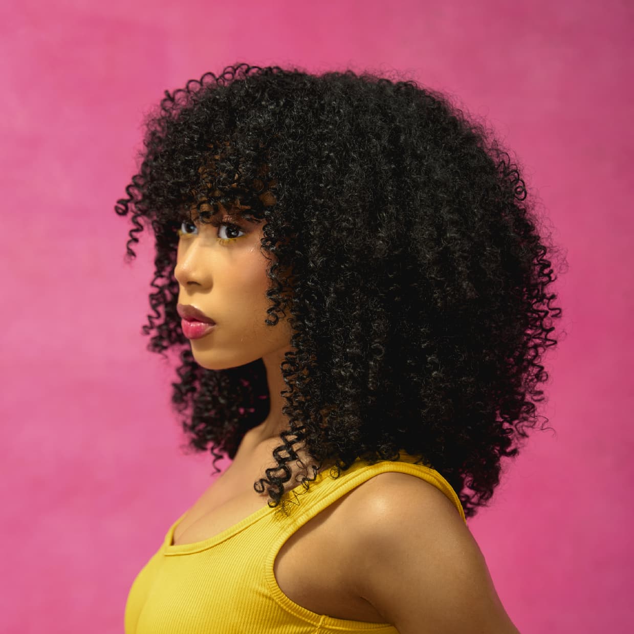 Side profile of a woman in the afro-curly and voluminous DIVA wig wearing a yellow top against a pink background