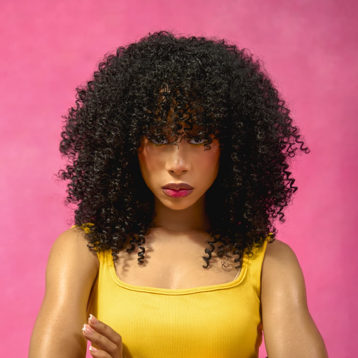 Woman in the diva wig wearing a yellow top against a pink background, she is staring straight into the camera