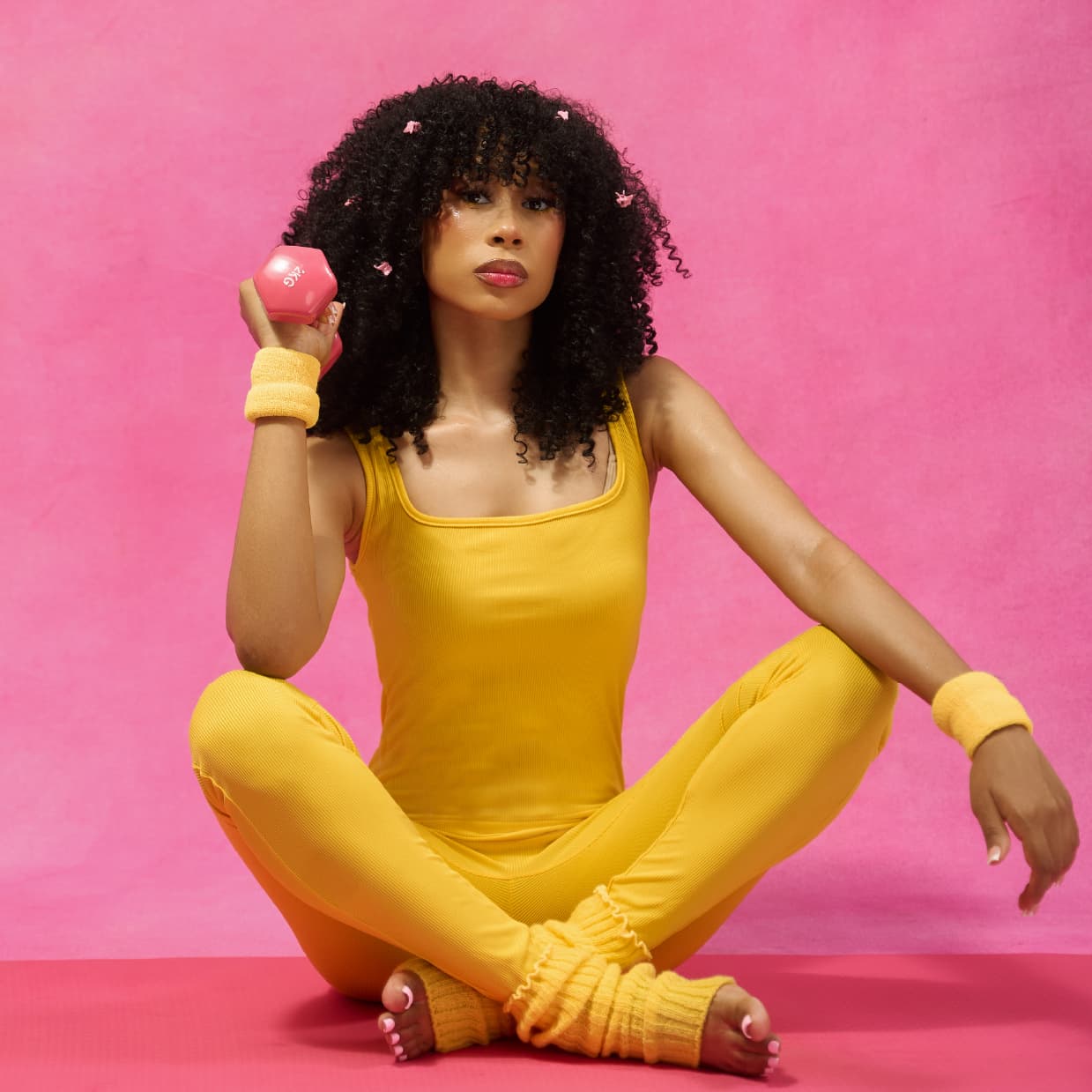 Woman in the Afro-curly and voluminous DIVA wig in a yellow outfit sitting on a pink background holding a pink dumbbell