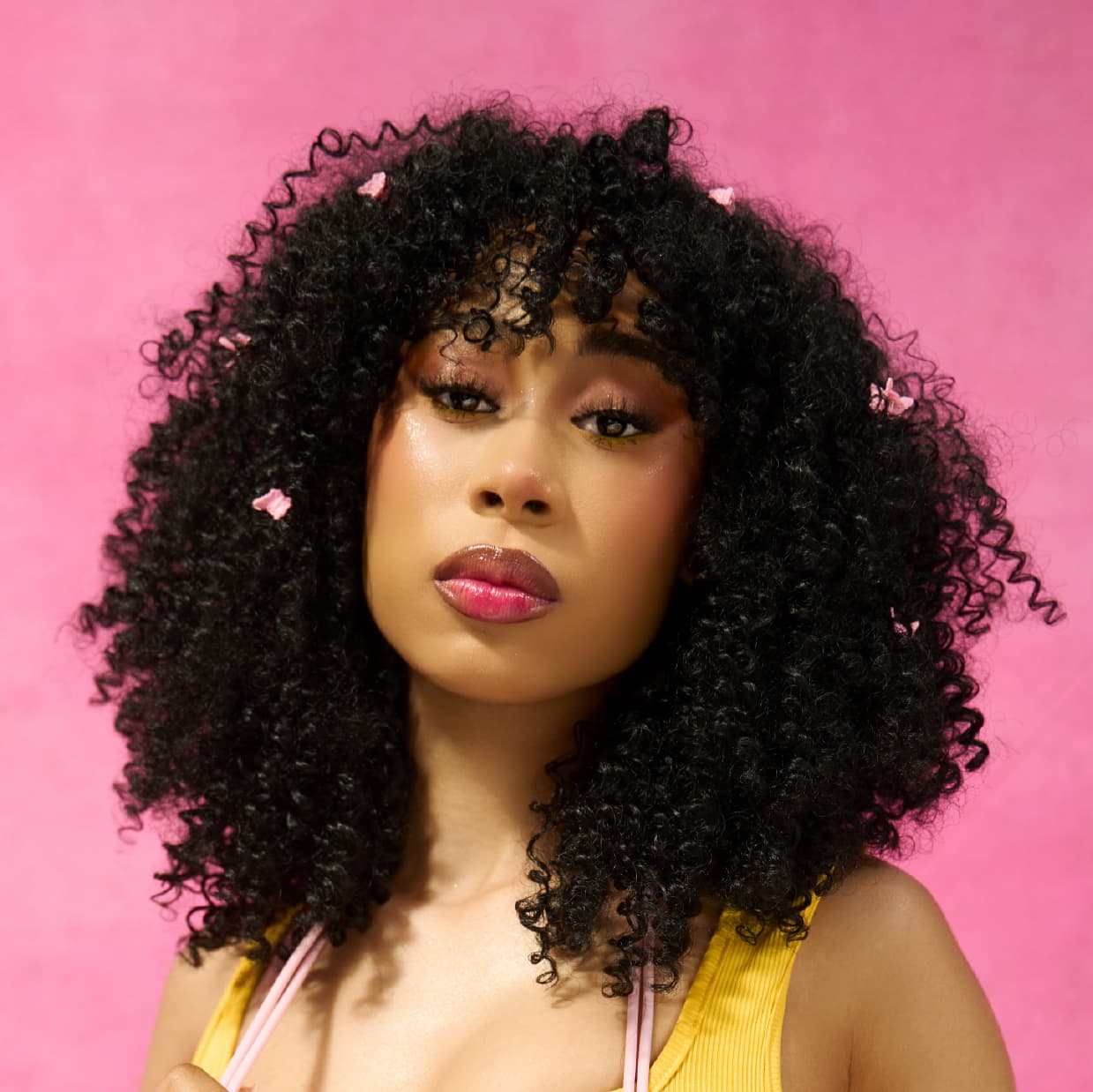 woman in the diva, an Afro curly voluminous wig, wearing a yellow top against a pink background