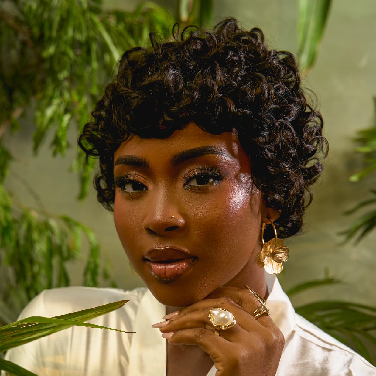 A woman wearing the muse wig (a short pixie hairstyle) and jewelry against a natural green background
