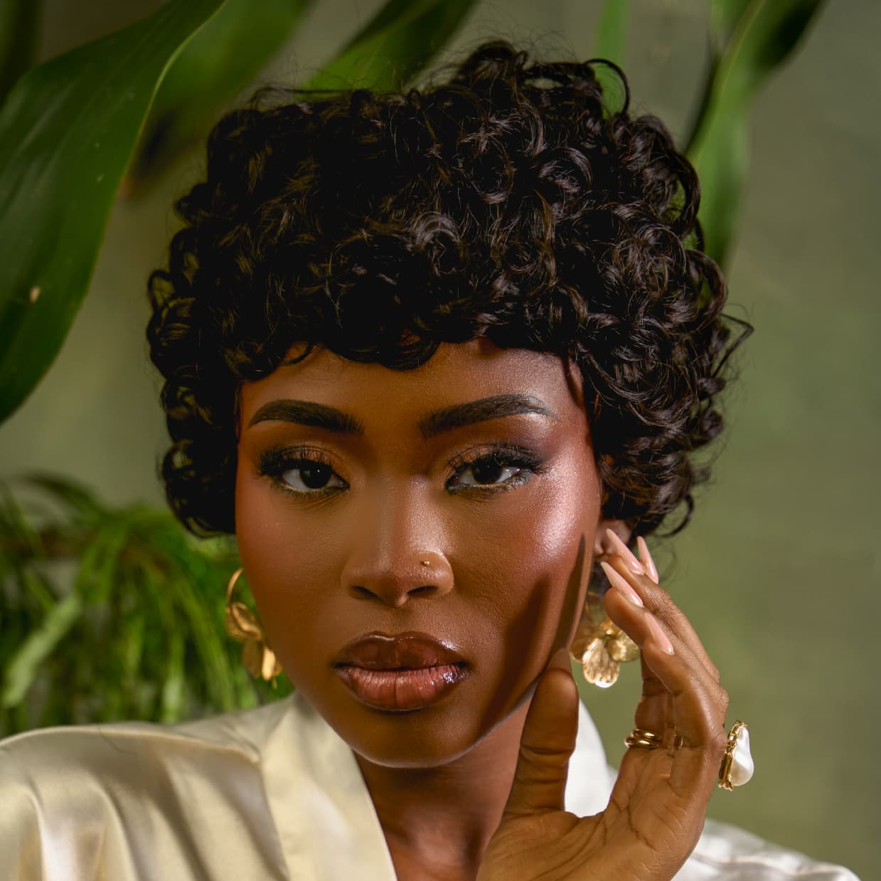 Woman in the Tress to Go short pixie wig called "The Muse" wearing gold earrings and rings, with a blurred green background