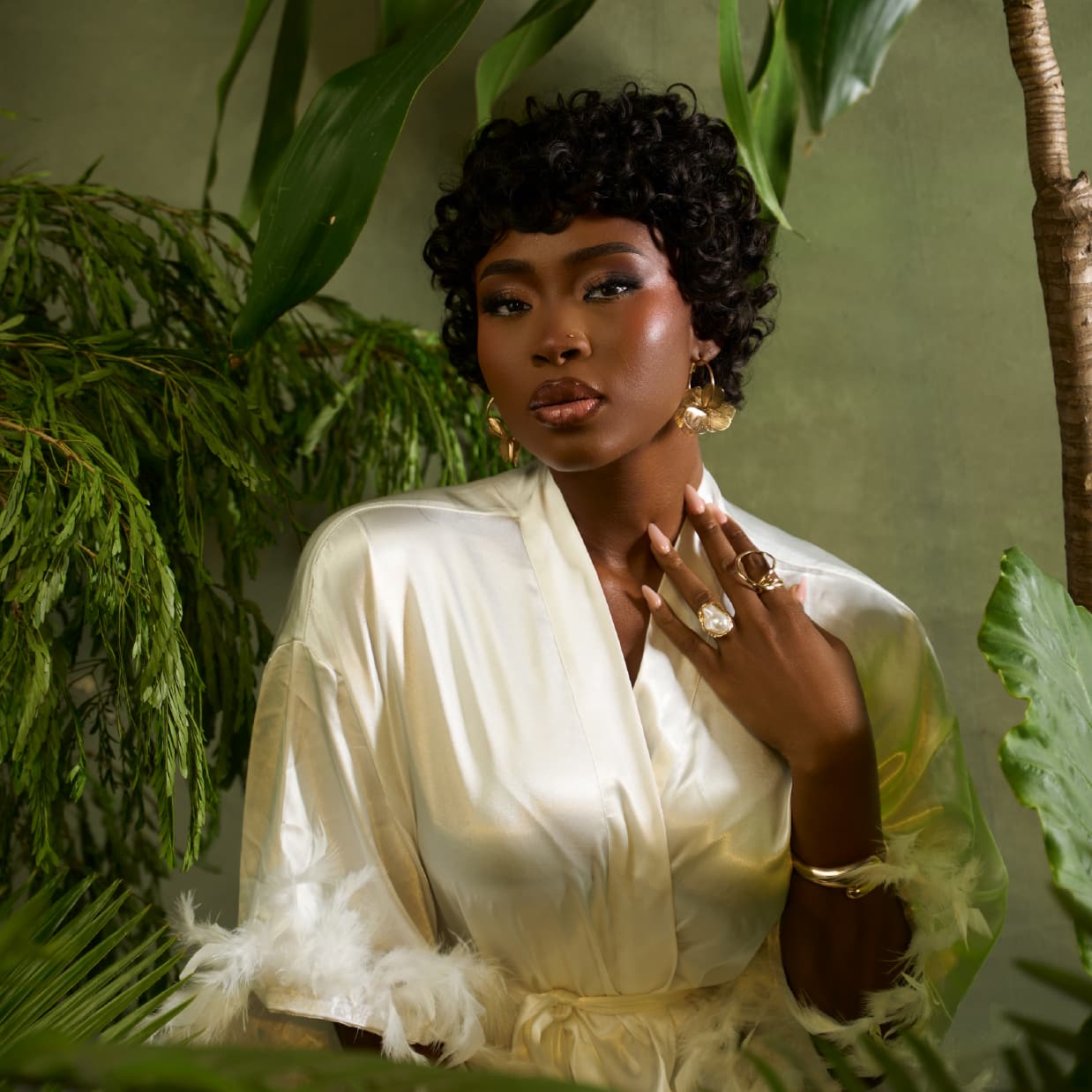 Woman in the Tress To Go pixie wig called the Muse wearing gold jewelry against a green leafy background