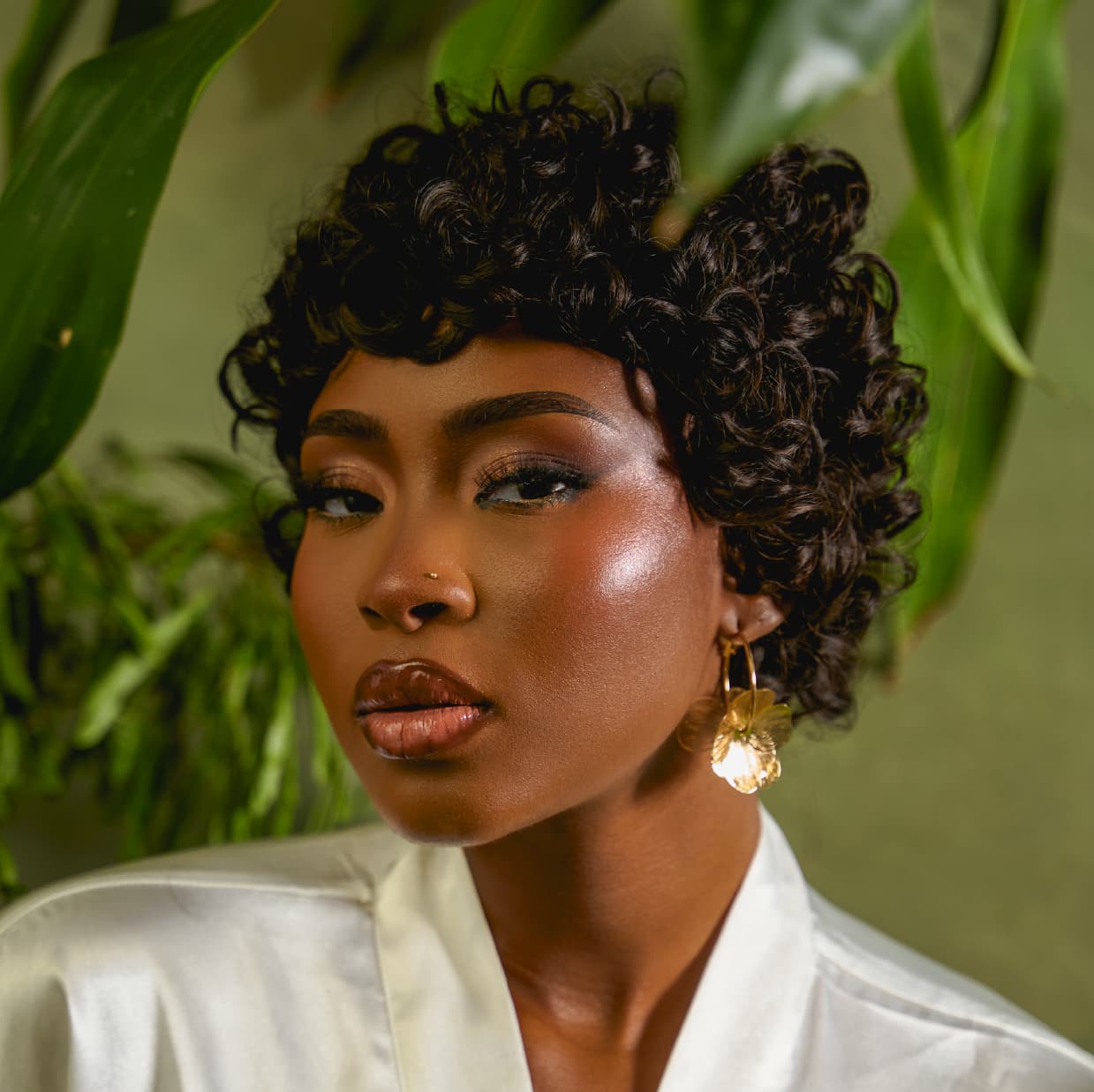 Woman in a Tress to Go pixie wig called "the muse" and earrings against a green leafy background